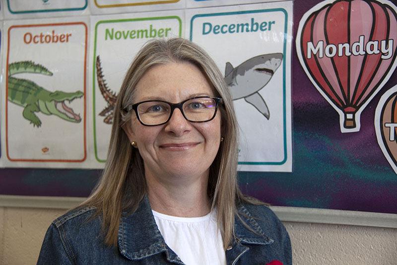 A portrait of Prace student Nikki who came to prace to study education support. She is smiling and standing in front of colourful educational posters.