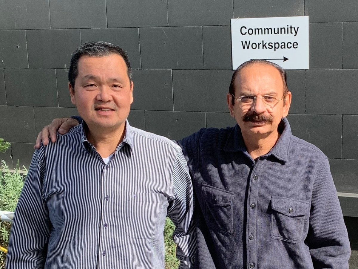 Tony is standing on the left and Fiaz is standing on the right with his arm around Tony. Both men wear collared long-sleeve shirts. Tony's is pinstripe with white and blue, and Fiaz's is dark blue. Fiaz is also wearing reading glasses. They are both standing in front of a brick wall painted dark grey.