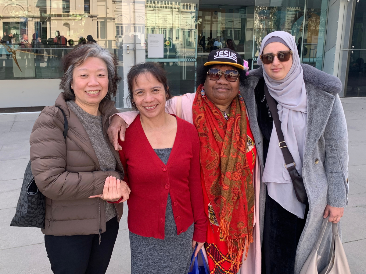 Four women from diverse backgrounds stand together smiling in front of the Melbourne Museum in Carlton, Victoria, Australia.